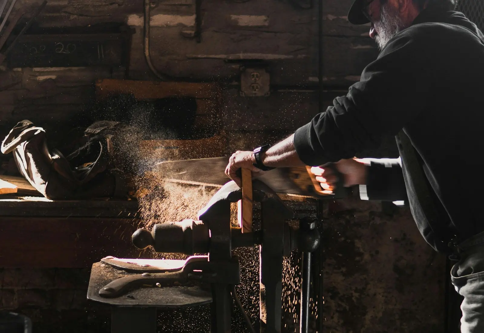 Un menuisier scie à la main une planche serrée dans l’étau, la poussière de bois en suspension dans un rai de lumière.